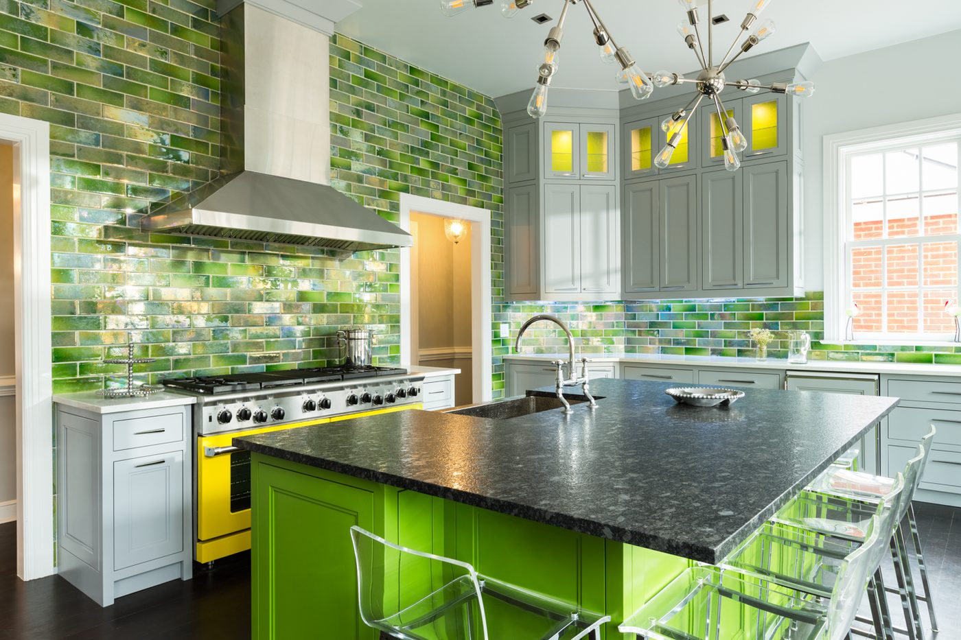 Bright interior photograph of a modern kitchen with green tile, gray cabinetry, and reflective metal fixtures